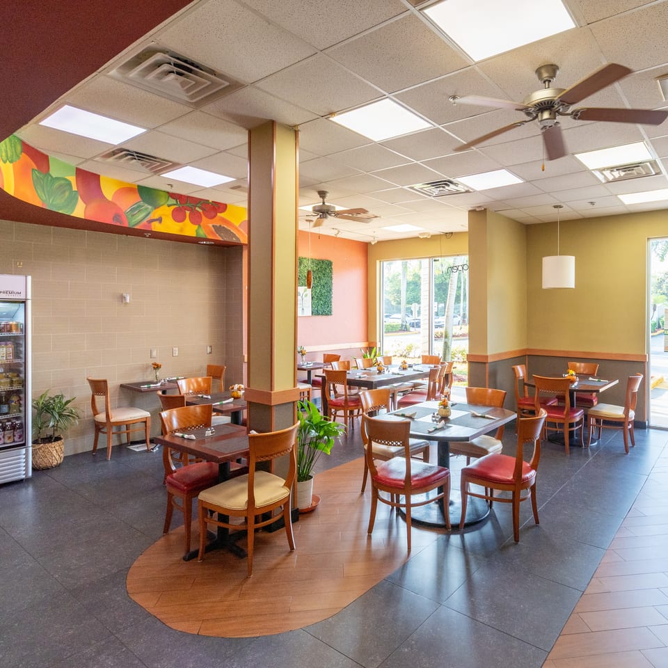 Bright restaurant dining room with neatly set tables, wooden chairs, colorful fruit mural, beverage refrigerator, plants, and ceiling fans.