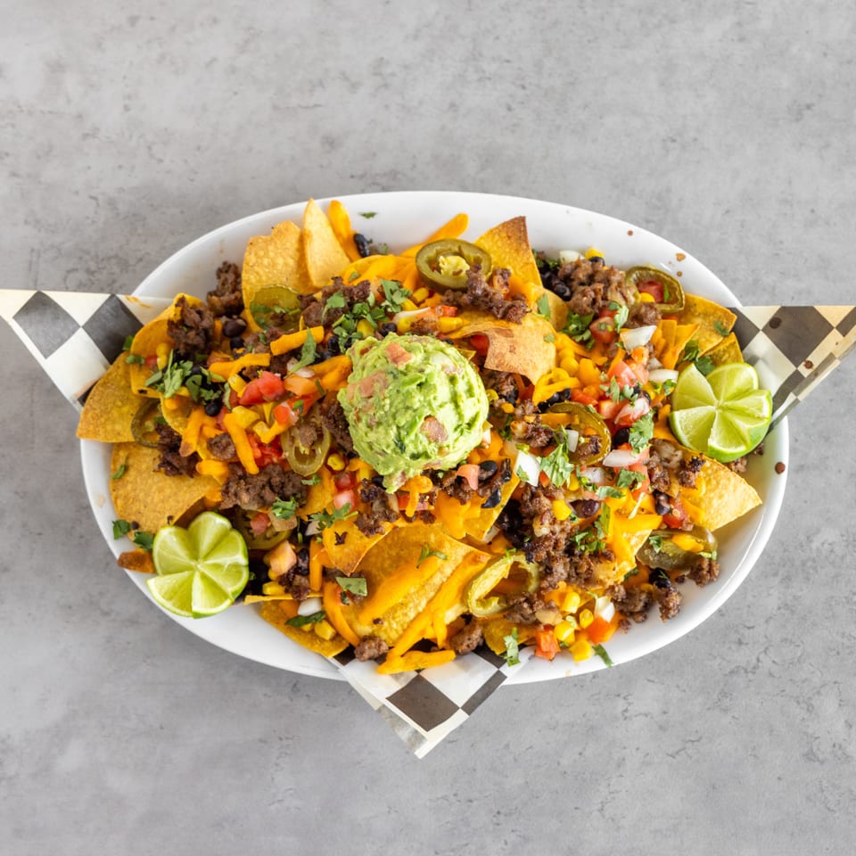 Loaded nachos topped with seasoned ground beef, melted cheese, guacamole, jalapeños, black beans, salsa, cilantro, and lime wedges.