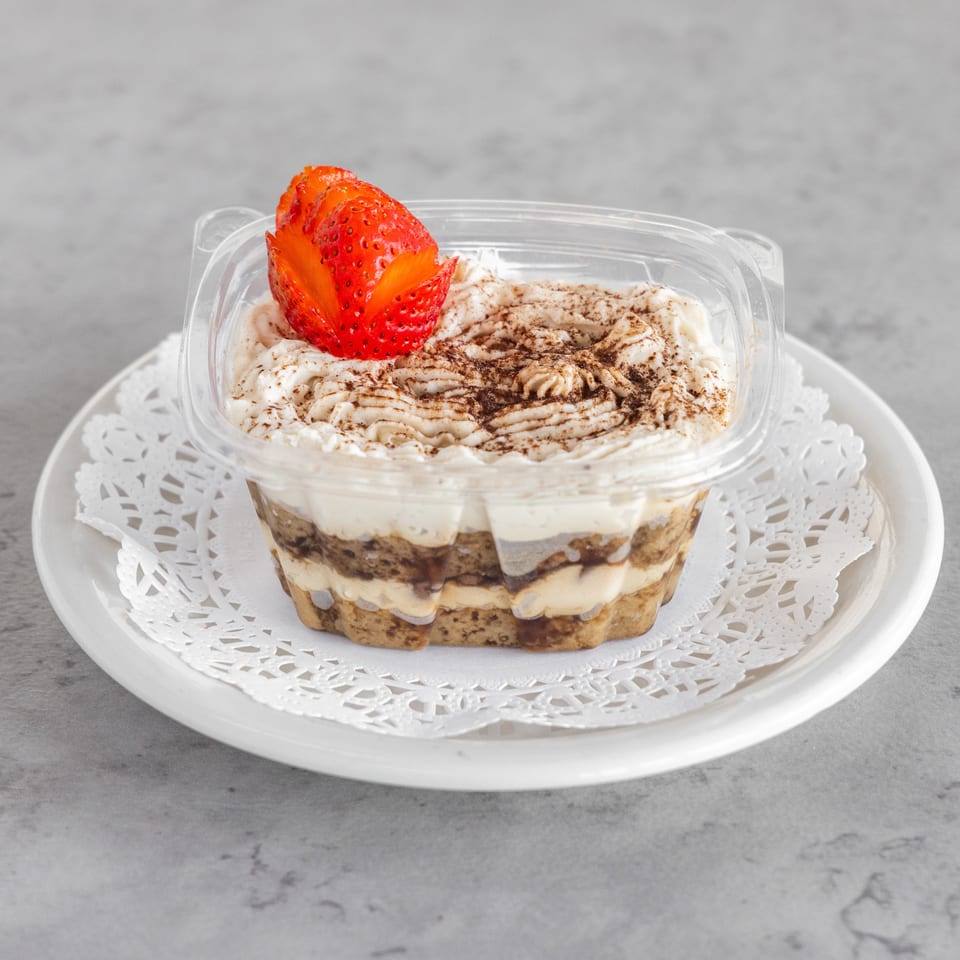 Layered tiramisu topped with whipped cream, cocoa dusting, and a carved strawberry, served in a clear container on a white plate.