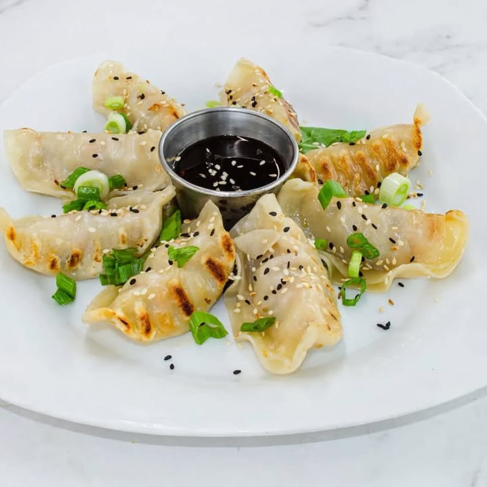 Pan-fried dumplings garnished with scallions and sesame seeds, arranged around a cup of dark dipping sauce on a white plate.