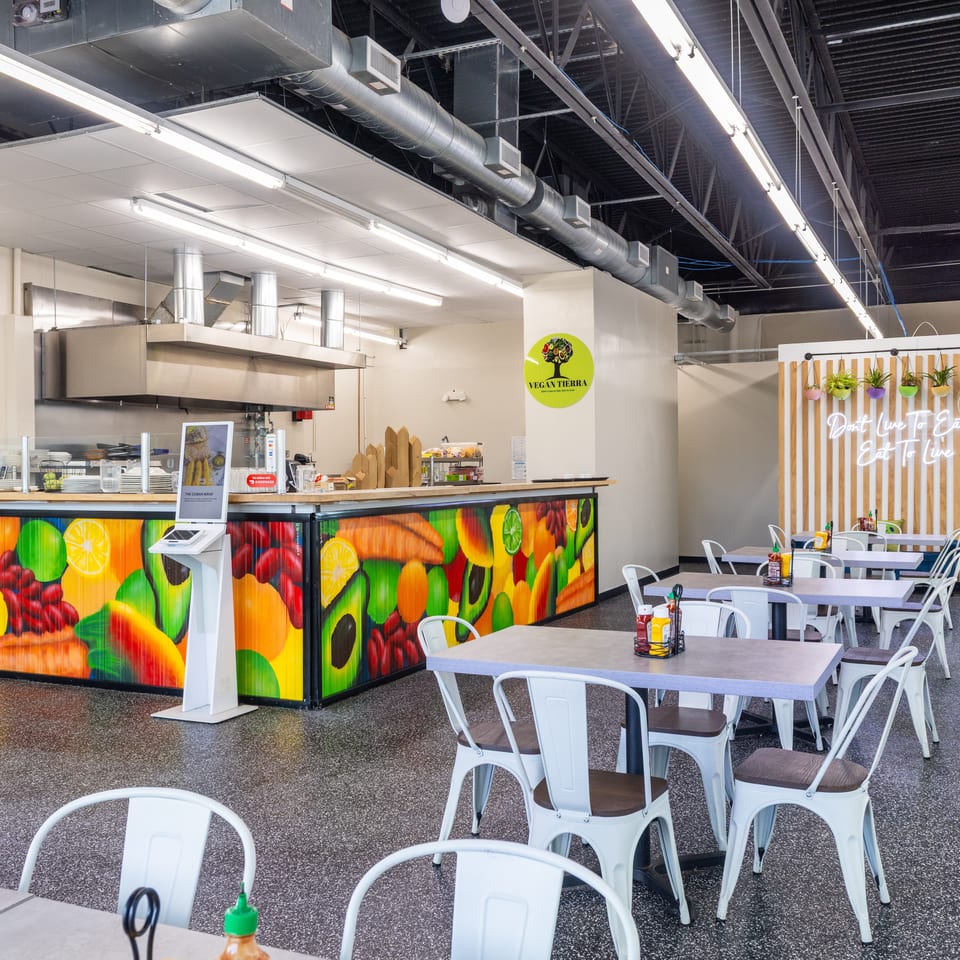 Bright Vegan Tierra restaurant interior with colorful fruit murals, open kitchen counter, white tables, chairs, and hanging plants.