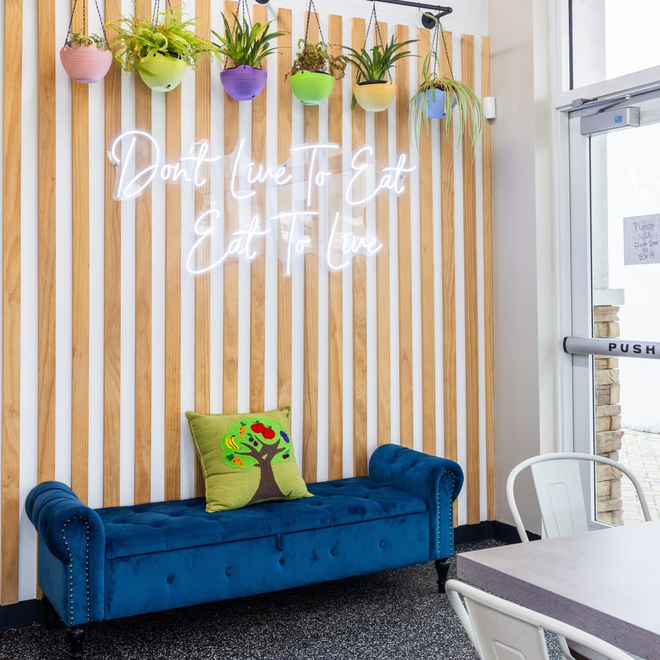 Colorful hanging plants and a glowing “Don’t Wait to Eat” sign decorate a wood-slat wall above a blue velvet sofa.