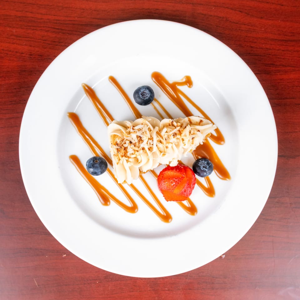 Slice of coconut-topped cake garnished with blueberries and strawberries on a white plate with caramel drizzle.