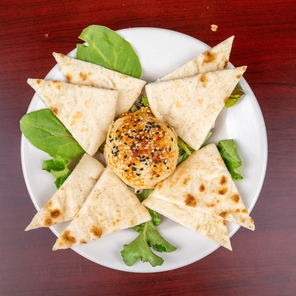 Overhead view of toasted flatbread wedges arranged around a sesame-topped baked dip on leafy greens.