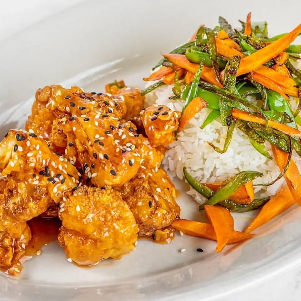 Sesame-glazed crispy chicken served with white rice and sautéed carrots and green peppers on a white plate.