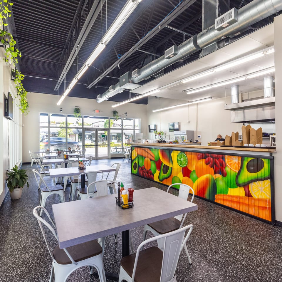 Bright restaurant dining room with white tables, metal chairs, hanging plants, exposed ducts, and a colorful fruit mural on the counter.