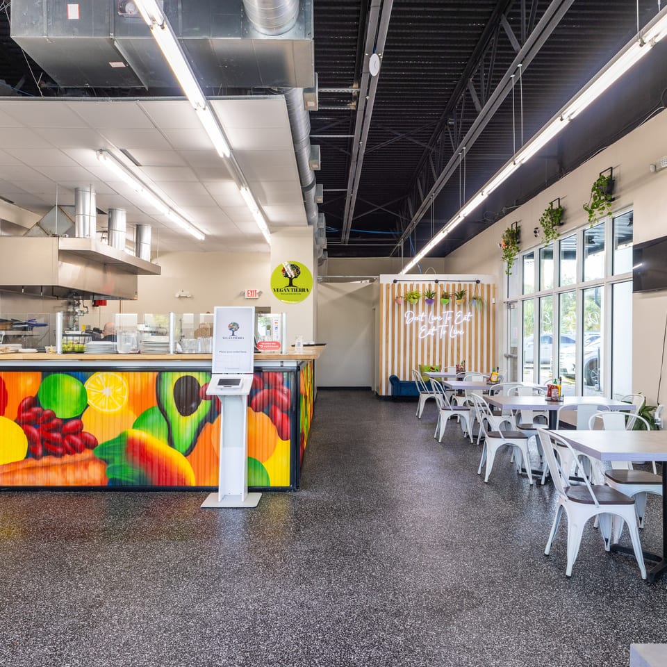 Bright Vegantierra restaurant interior with colorful fruit mural, open kitchen, white seating, hanging plants, and large windows.
