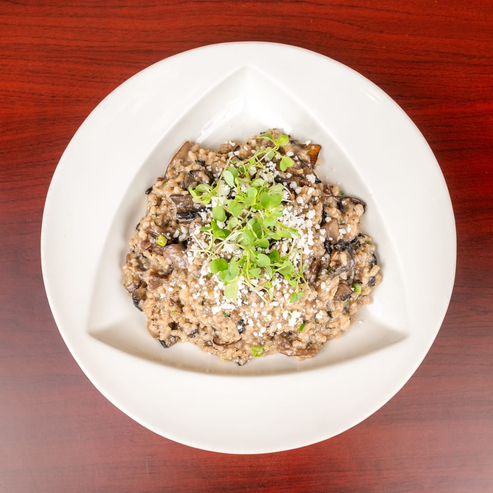 Creamy mushroom risotto topped with crumbled cheese and microgreens on a white plate over a wooden table.