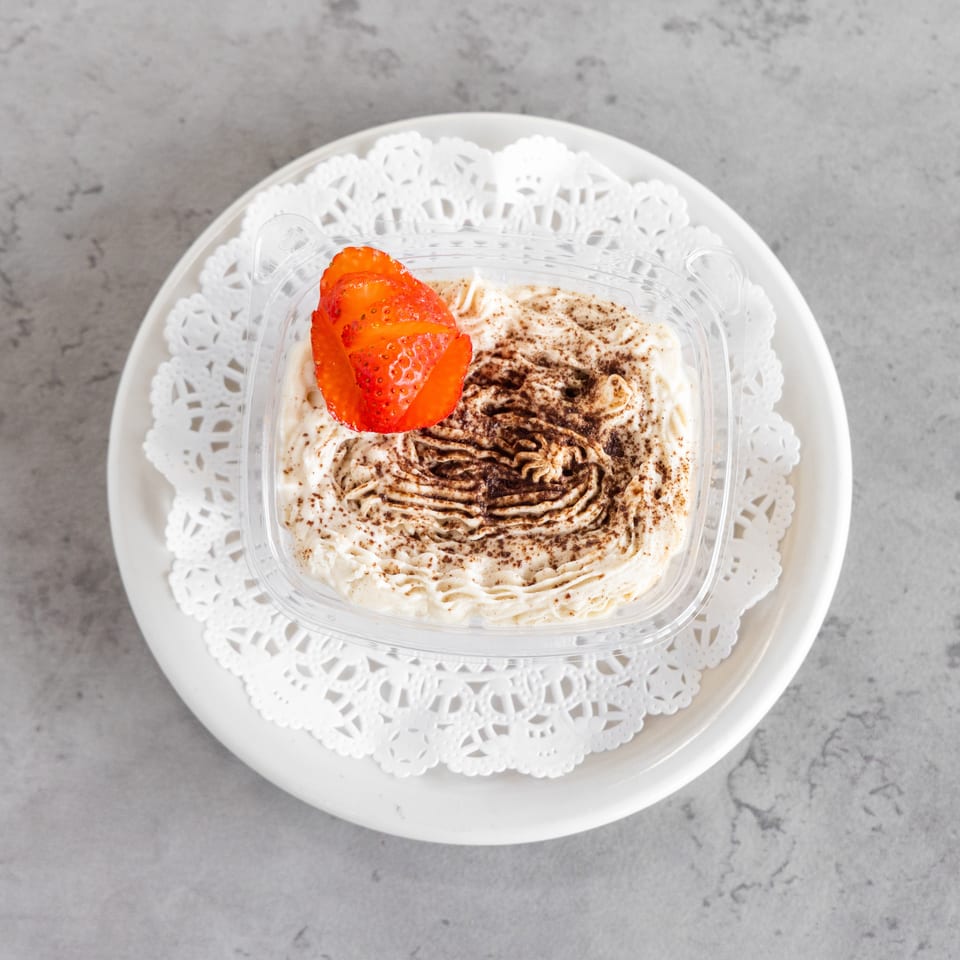 Creamy tiramisu topped with cocoa powder and sliced strawberry in a clear container on a decorative white plate.