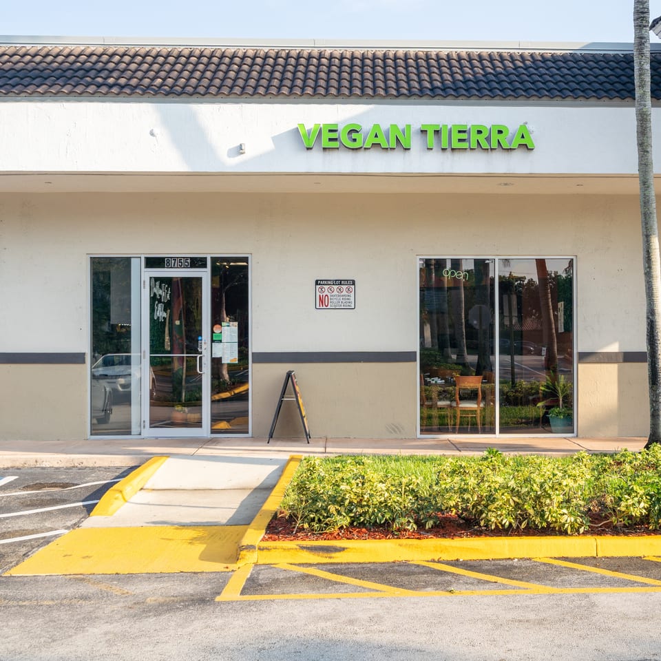 Exterior of Vegan Tierra restaurant with green signage, glass entrance, tiled roof, palm tree, and landscaped storefront.