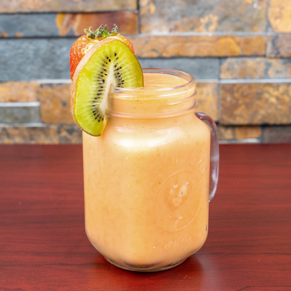 Creamy orange smoothie in a glass jar, garnished with sliced kiwi and strawberry, on a wooden table against a tiled wall.