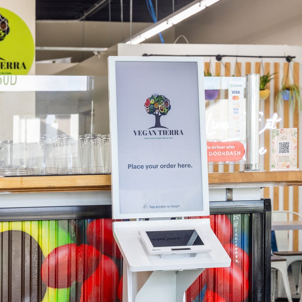 Vegan Tierra restaurant counter with a self-service ordering kiosk, glassware, payment sign, and colorful produce-themed decor.