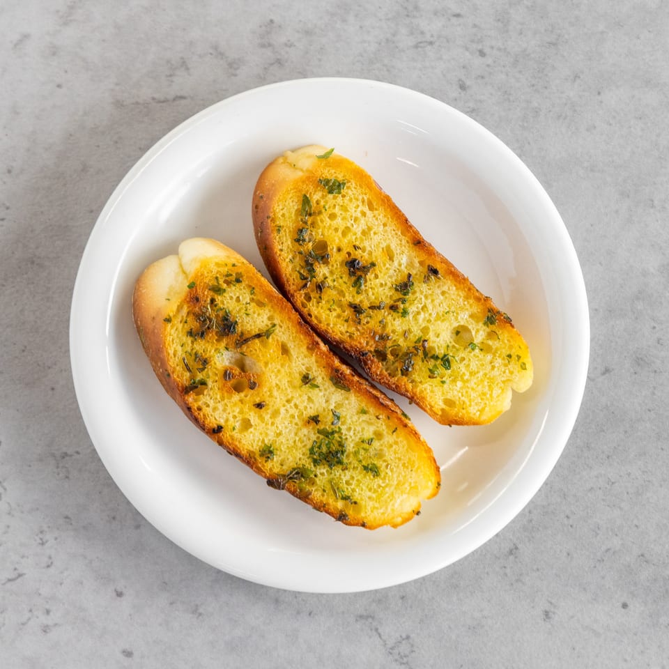 Two slices of golden garlic herb toast arranged on a white plate over a gray countertop.