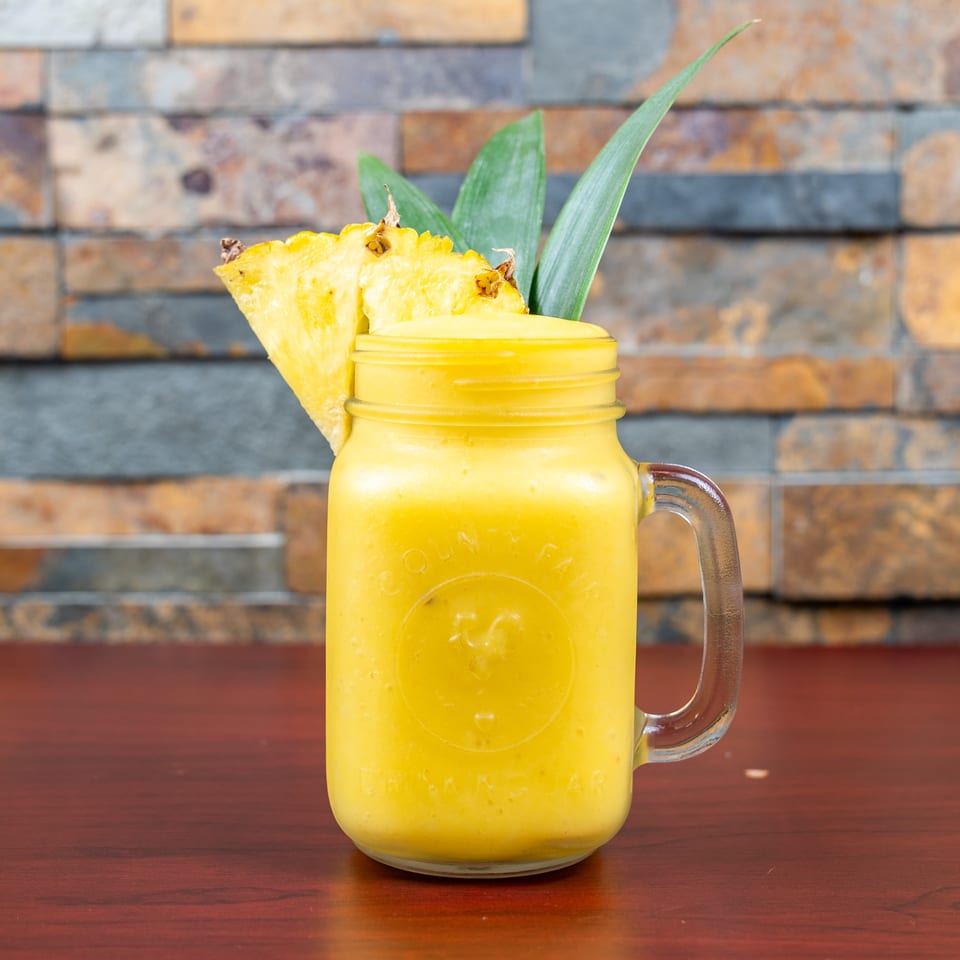 Bright yellow pineapple smoothie in a glass mug, garnished with pineapple slices and leaves on a wooden table.