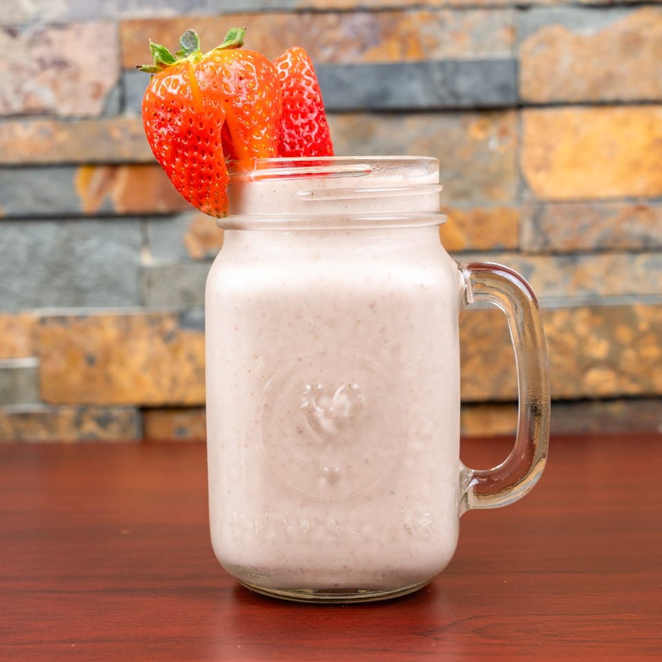 Creamy strawberry smoothie in a glass mug, garnished with fresh strawberries, sits on a wooden table against a brick wall.
