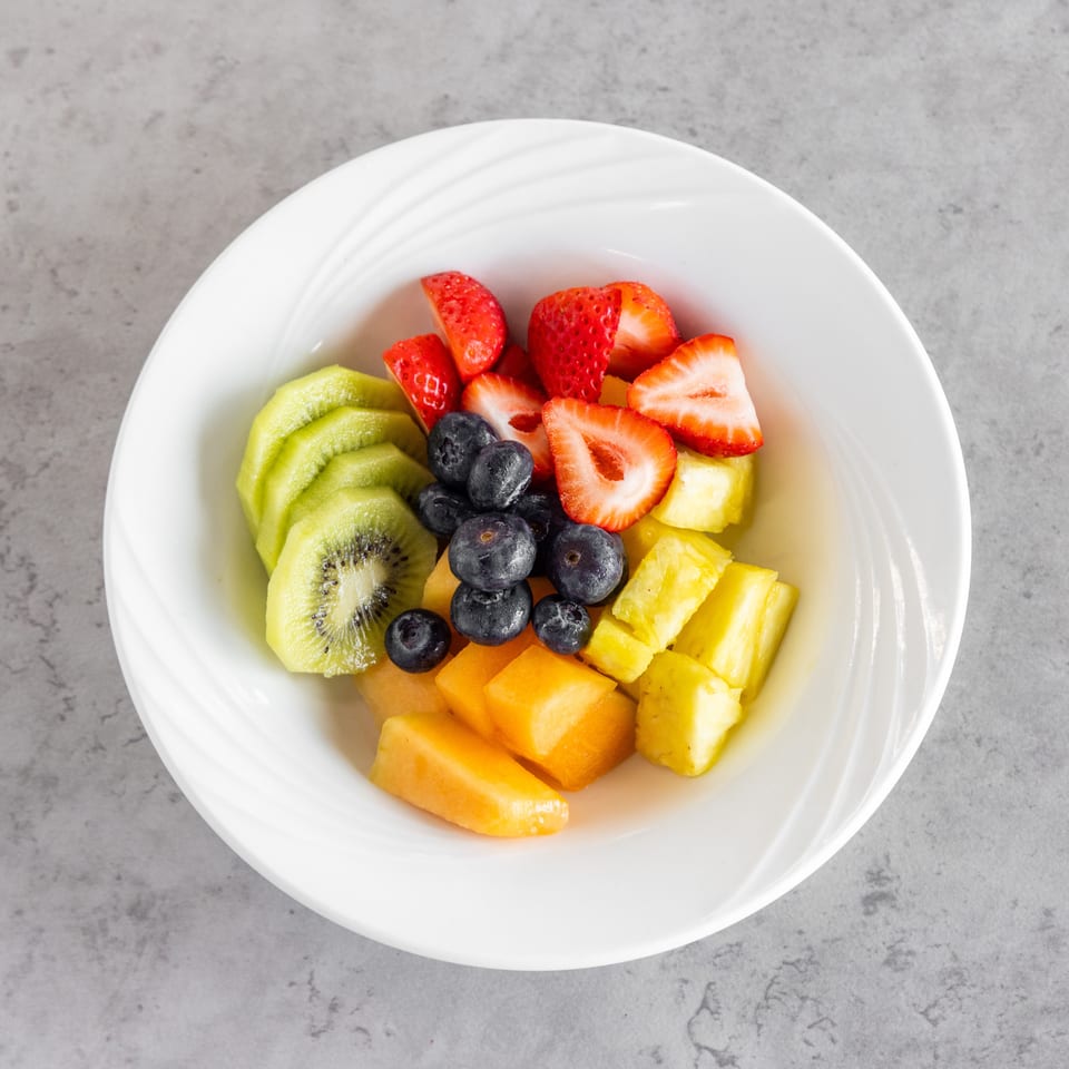 Bowl of sliced kiwi, strawberries, blueberries, pineapple, and cantaloupe on a white plate.