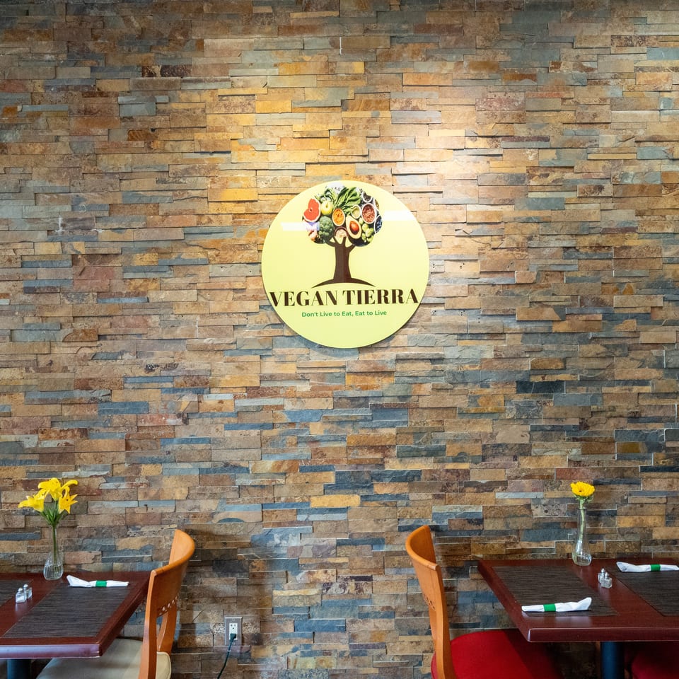 Vegan Tierra restaurant dining area with a colorful food-themed logo on a stone accent wall, tables, chairs, and yellow flowers.