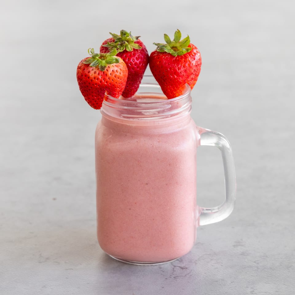 Pink strawberry smoothie in a clear glass mug, topped with three fresh strawberries on a light countertop.