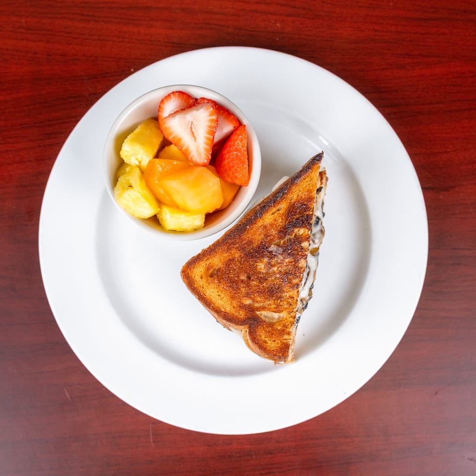 Grilled cheese sandwich served with a bowl of pineapple, mango, orange, and strawberry fruit on a white plate.