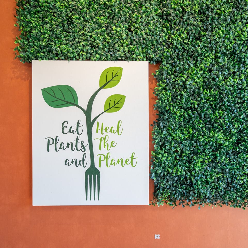 Green leafy wall surrounds a sign reading “Eat Plants and Heal the Planet,” featuring a fork-shaped plant illustration.