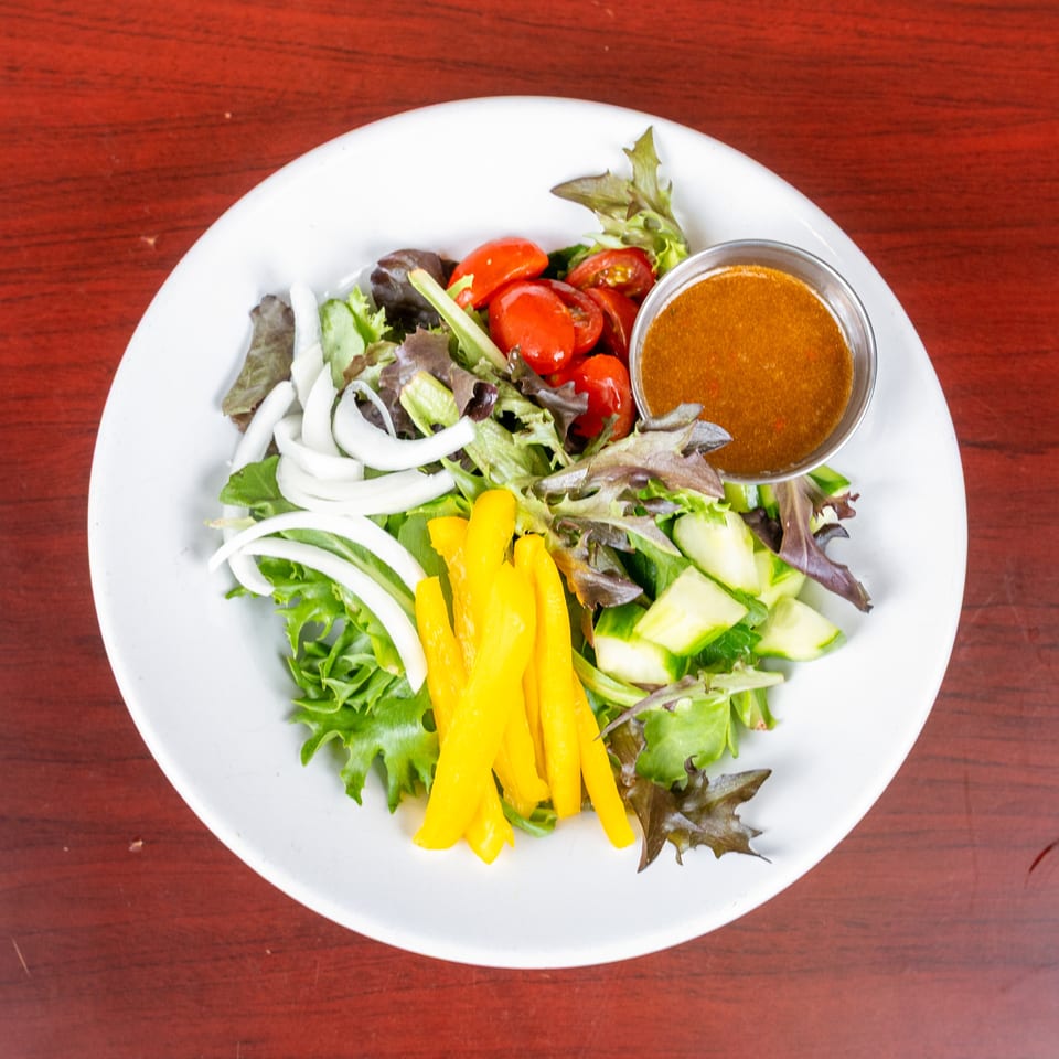 Mixed green salad with tomatoes, onions, cucumber, yellow peppers, and dressing on a white plate atop a wooden table.