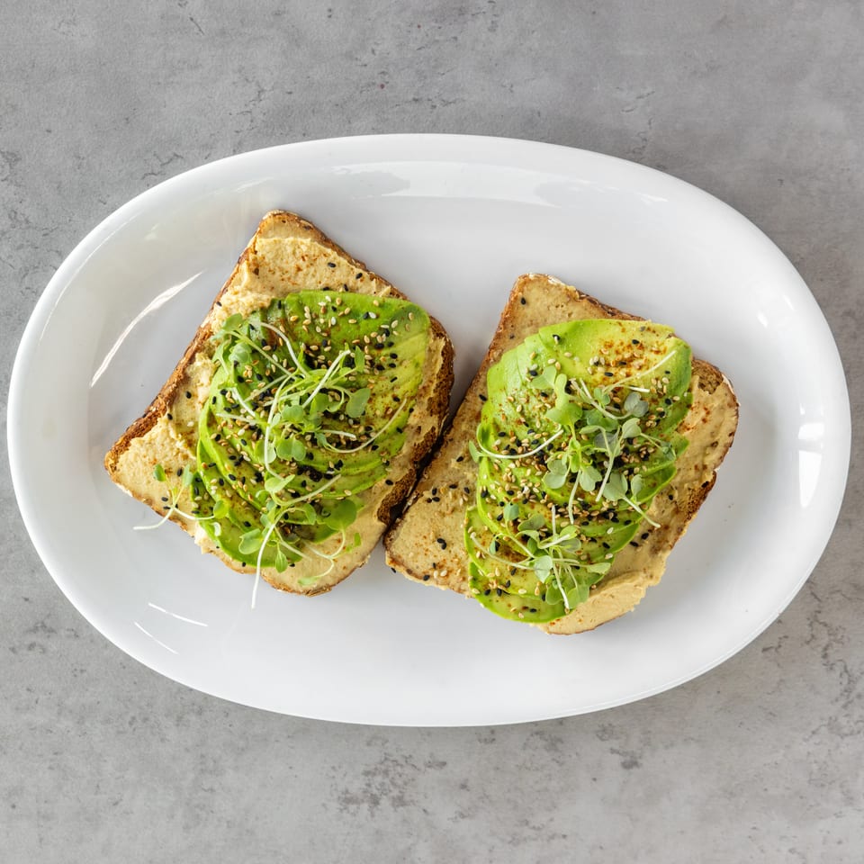 Two slices of avocado toast topped with hummus, microgreens, and sesame seeds on a white plate.