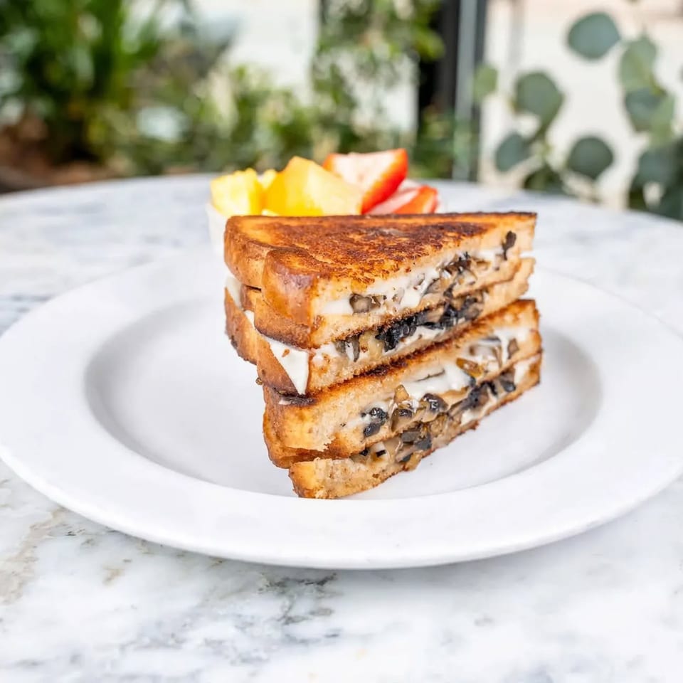 Grilled mushroom and cheese sandwich stacked on a white plate with sliced fruit, set on a bright marble table.