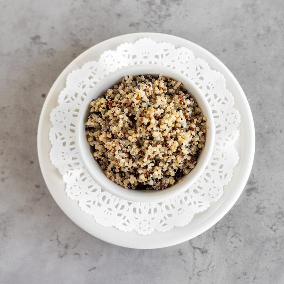 Bowl of tri-color quinoa served on a decorative white doily and plate atop a gray stone surface.