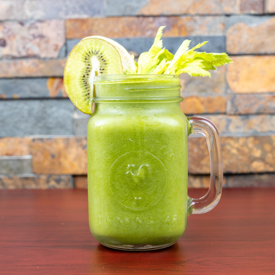Green smoothie in a glass mason jar garnished with kiwi and celery, set on a wooden table against a brick wall.